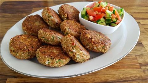 A white plate holding several golden-brown falafel patties arranged in a semicircle, with a small bowl of chopped salad consisting of tomatoes, cucumbers, and green peppers on the side. The plate is set on a wooden tabletop.