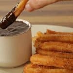 A plate of churros is served next to a white cup filled with thick chocolate sauce. A hand is dipping one churro into the chocolate sauce. The background is a wooden table.