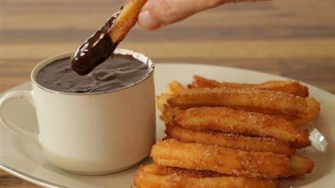 A plate of churros is served next to a white cup filled with thick chocolate sauce. A hand is dipping one churro into the chocolate sauce. The background is a wooden table.