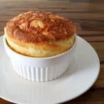 A golden-brown soufflé rises impressively out of a white ramekin, placed on a matching white saucer. The background features a wooden surface with natural grain patterns, adding a warm, rustic touch to the image.