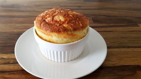 A golden-brown soufflé rises impressively out of a white ramekin, placed on a matching white saucer. The background features a wooden surface with natural grain patterns, adding a warm, rustic touch to the image.