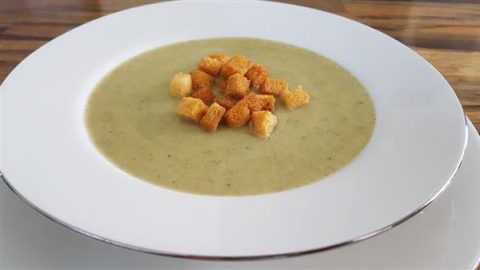 A white plate filled with creamy broccoli soup, garnished with a cluster of golden-brown croutons in the center, placed on a wooden table. The soup has a smooth texture, while the croutons add a contrasting crispness.