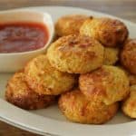A white plate featuring a stack of golden-brown, crispy cauliflower tots is placed on a wooden table. A small, square white bowl filled with red dipping sauce accompanies the tots on the plate. The tots appear well-baked and appetizing.
