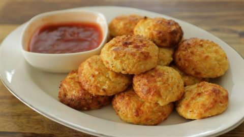 A white plate featuring a stack of golden-brown, crispy cauliflower tots is placed on a wooden table. A small, square white bowl filled with red dipping sauce accompanies the tots on the plate. The tots appear well-baked and appetizing.