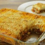 A glass baking dish filled with baked shepherd's pie, showing a layer of golden-brown mashed potatoes on top with visible ground meat and vegetables underneath. A piece has been cut out and sits on a white plate in the background.