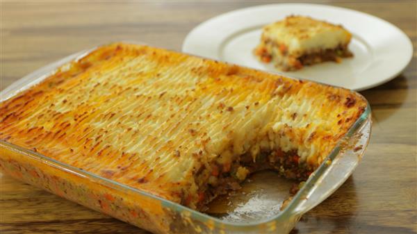 A glass baking dish filled with baked shepherd's pie, showing a layer of golden-brown mashed potatoes on top with visible ground meat and vegetables underneath. A piece has been cut out and sits on a white plate in the background.