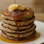 A stack of six pancakes topped with sliced bananas and drizzled with syrup on a white plate. The syrup is flowing down the sides of the pancakes. The background is a wooden table.