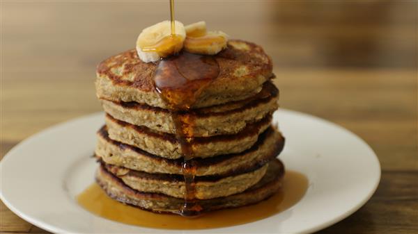 A stack of six pancakes topped with sliced bananas and drizzled with syrup on a white plate. The syrup is flowing down the sides of the pancakes. The background is a wooden table.