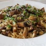 A close-up of a plate of mushroom risotto garnished with chopped parsley. The creamy rice dish is served on a white, decorative plate and placed on a wooden table. The risotto is a mixture of rice, mushrooms, and herbs, giving it a rich and savory appearance.