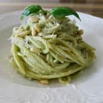 A plate of spaghetti with creamy green pesto sauce, garnished with pine nuts, grated cheese, and a sprig of fresh basil, presented on a white decorative plate. The background shows a wooden table.