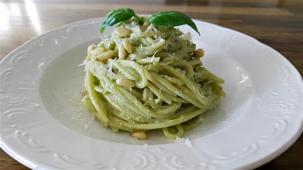 A plate of spaghetti with creamy green pesto sauce, garnished with pine nuts, grated cheese, and a sprig of fresh basil, presented on a white decorative plate. The background shows a wooden table.