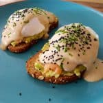 A vibrant blue plate holds two pieces of toast topped with mashed avocado, poached eggs, creamy hollandaise sauce, black sesame seeds, and finely chopped chives. The dish is artistically plated on a wooden tabletop.