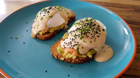 A vibrant blue plate holds two pieces of toast topped with mashed avocado, poached eggs, creamy hollandaise sauce, black sesame seeds, and finely chopped chives. The dish is artistically plated on a wooden tabletop.