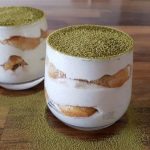 Two glass cups of matcha tiramisu sit on a wooden surface. The dessert features layers of creamy mascarpone and soft ladyfinger biscuits, topped with a dusting of vibrant green matcha powder. Some matcha powder is also sprinkled around the base of the cups.