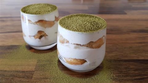 Two glass cups of matcha tiramisu sit on a wooden surface. The dessert features layers of creamy mascarpone and soft ladyfinger biscuits, topped with a dusting of vibrant green matcha powder. Some matcha powder is also sprinkled around the base of the cups.