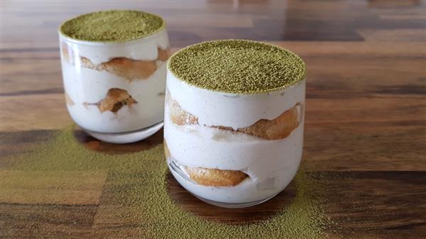 Two glass cups of matcha tiramisu sit on a wooden surface. The dessert features layers of creamy mascarpone and soft ladyfinger biscuits, topped with a dusting of vibrant green matcha powder. Some matcha powder is also sprinkled around the base of the cups.