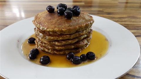 A stack of pancakes topped with blueberries and drizzled with maple syrup, served on a white plate on a wooden table. The syrup pools around the base of the pancakes, with extra blueberries scattered on the plate.