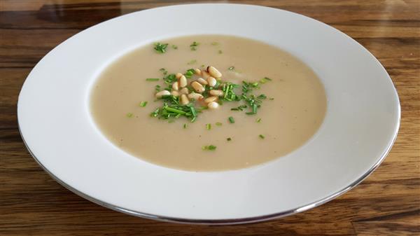 A white bowl filled with creamy soup, garnished with chopped chives and pine nuts, sits on a wooden table.