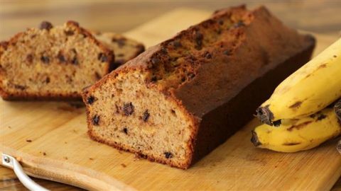 A sliced loaf of banana bread with chocolate chips on a wooden cutting board. A few ripe bananas are placed next to the bread. The interior of the bread is moist and speckled with chocolate chips, while the crust appears slightly browned.