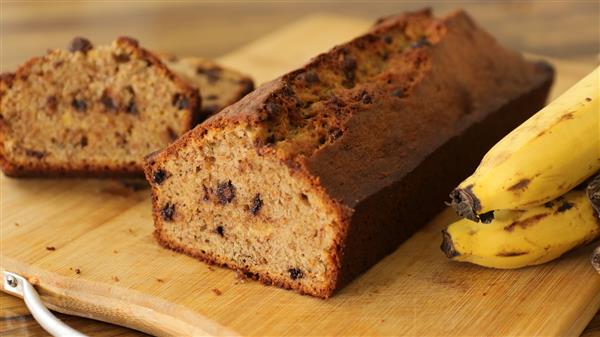 A sliced loaf of banana bread with chocolate chips on a wooden cutting board. A few ripe bananas are placed next to the bread. The interior of the bread is moist and speckled with chocolate chips, while the crust appears slightly browned.