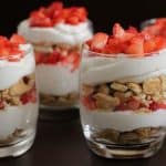 Four glass cups filled with layered desserts, consisting of whipped cream, crushed cookies, and chopped strawberries. The layers are neatly arranged, creating a visually appealing contrast of red, white, and beige tones.