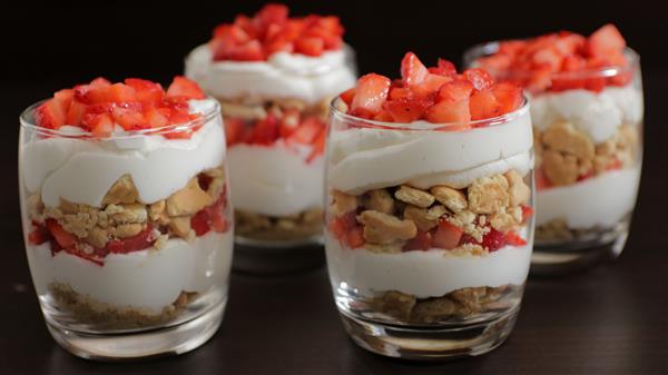 Four glass cups filled with layered desserts, consisting of whipped cream, crushed cookies, and chopped strawberries. The layers are neatly arranged, creating a visually appealing contrast of red, white, and beige tones.