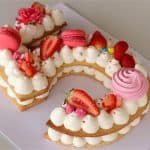 A cake shaped like the number 2 is decorated with layers of cream, pink macarons, strawberries, meringues, and small flowers on a white surface. The cake features vibrant red, pink, and white colors.