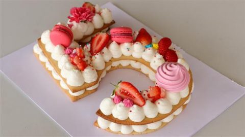 A cake shaped like the number 2 is decorated with layers of cream, pink macarons, strawberries, meringues, and small flowers on a white surface. The cake features vibrant red, pink, and white colors.