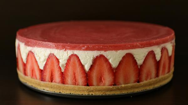 A round, layered strawberry cheesecake with a golden graham cracker crust, a middle layer of thick, creamy white filling, and topped with a smooth, red strawberry layer. Fresh strawberry slices line the sides between the crust and the top layer.