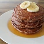 A stack of pancakes topped with banana slices and drizzled with syrup sits on a white plate. The plate is set on a wooden table.