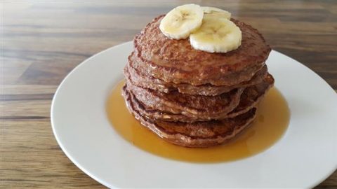 A stack of pancakes topped with banana slices and drizzled with syrup sits on a white plate. The plate is set on a wooden table.