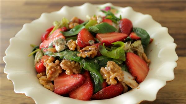 A white fluted bowl is filled with a fresh salad consisting of spinach leaves, sliced strawberries, walnuts, and bits of cheese. The salad is vibrant and colorful, placed on a wooden surface.