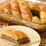A glass baking dish filled with sliced pieces of golden-brown baklava sits on a wooden table. A triangular piece of baklava, garnished with crushed nuts, is served on a white plate in the foreground, accompanied by a small gold fork.
