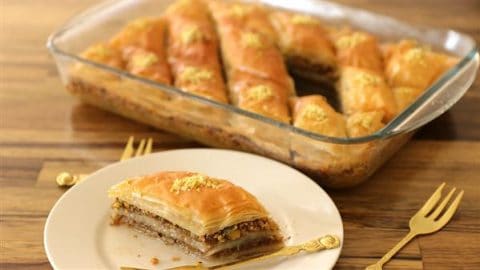 A glass baking dish filled with sliced pieces of golden-brown baklava sits on a wooden table. A triangular piece of baklava, garnished with crushed nuts, is served on a white plate in the foreground, accompanied by a small gold fork.