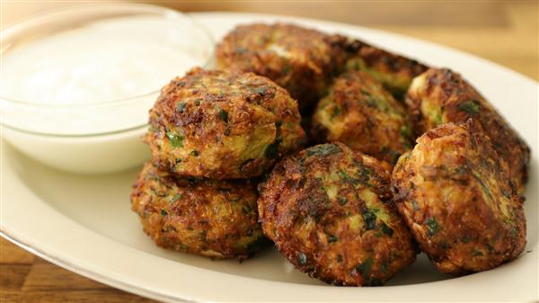 A plate of golden-brown fritters garnished with herbs, served alongside a small bowl of white dipping sauce. The fritters appear crispy and well-seasoned. The background is a blurred wooden surface.