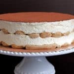 A traditional Italian tiramisu dessert sits on a white cake stand. It features layers of coffee-soaked ladyfingers and creamy mascarpone, topped with a dusting of cocoa powder. The background is dark, highlighting the cake's rich textures.