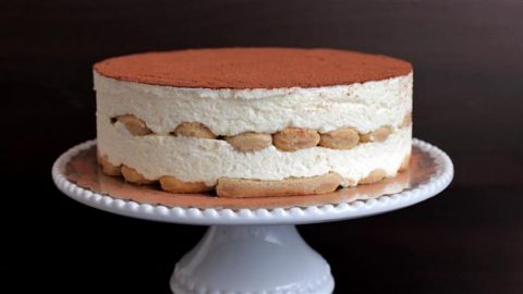 A traditional Italian tiramisu dessert sits on a white cake stand. It features layers of coffee-soaked ladyfingers and creamy mascarpone, topped with a dusting of cocoa powder. The background is dark, highlighting the cake's rich textures.