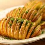 A white plate with three golden-brown Hasselback potatoes garnished with chopped green herbs and a sprinkle of black pepper. The potatoes are thinly sliced and slightly fanned out, revealing a crispy texture. The background is a wooden table surface.