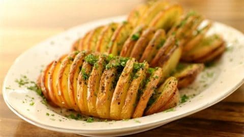 A white plate with three golden-brown Hasselback potatoes garnished with chopped green herbs and a sprinkle of black pepper. The potatoes are thinly sliced and slightly fanned out, revealing a crispy texture. The background is a wooden table surface.