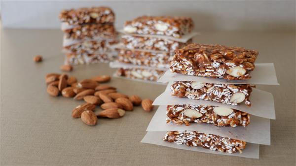 A close-up of several granola bars stacked on a surface, each separated by parchment paper. In the background, more granola bars are stacked, with scattered almonds nearby. The bars contain visible oats, nuts, and seeds.