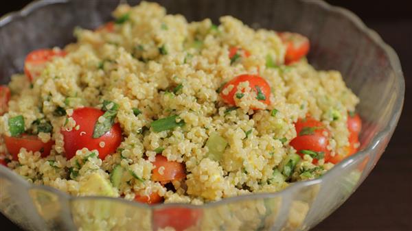 A clear glass bowl filled with a quinoa salad. The salad includes quinoa, cherry tomato halves, chopped cucumber, and fresh herbs like parsley or cilantro. The ingredients are mixed together, showcasing a colorful and fresh appearance.