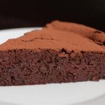 A close-up of a slice of flourless chocolate cake on a white plate. The cake is dense and rich, with a slightly cracked, dry-looking top and a moist, fudgy interior. The background is blurred and dark, making the cake stand out.