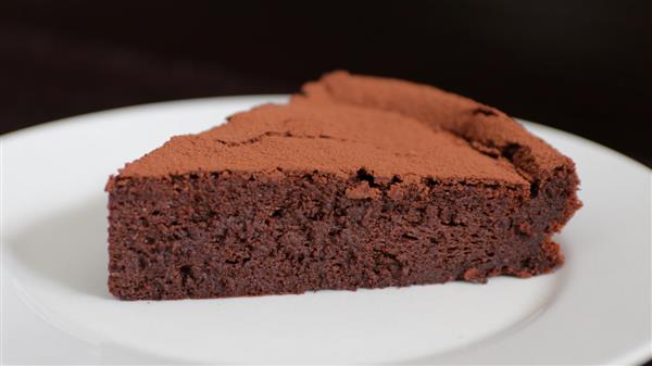 A close-up of a slice of flourless chocolate cake on a white plate. The cake is dense and rich, with a slightly cracked, dry-looking top and a moist, fudgy interior. The background is blurred and dark, making the cake stand out.