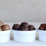 Three white bowls, each containing an array of energy bites or protein balls. The bites in each bowl have different colors and textures, suggesting a variety of ingredients and flavors. The bowls are placed in a row on a light-colored surface against a neutral background.