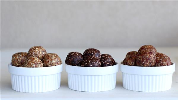 Three white bowls, each containing an array of energy bites or protein balls. The bites in each bowl have different colors and textures, suggesting a variety of ingredients and flavors. The bowls are placed in a row on a light-colored surface against a neutral background.