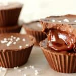 Close-up of chocolate cups filled with caramel, topped with a sprinkle of sea salt. One of the cups is bitten into, revealing the gooey caramel center. Additional whole chocolate cups are stacked in the background, all placed on a cream-colored surface.