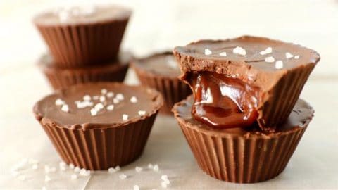 Close-up of chocolate cups filled with caramel, topped with a sprinkle of sea salt. One of the cups is bitten into, revealing the gooey caramel center. Additional whole chocolate cups are stacked in the background, all placed on a cream-colored surface.