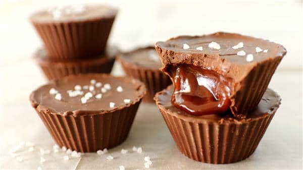 Close-up of chocolate cups filled with caramel, topped with a sprinkle of sea salt. One of the cups is bitten into, revealing the gooey caramel center. Additional whole chocolate cups are stacked in the background, all placed on a cream-colored surface.