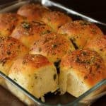 A glass baking dish filled with freshly baked herb dinner rolls. The rolls are golden brown on top with visible flecks of green herbs, indicating a savory seasoning. One roll in the front appears to have been partially broken open, displaying a soft, fluffy interior.