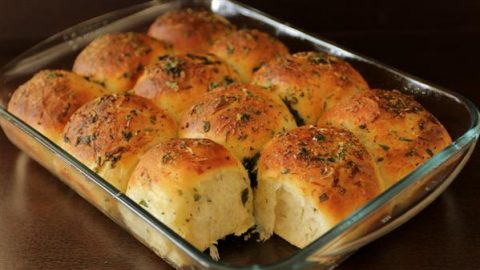 A glass baking dish filled with freshly baked herb dinner rolls. The rolls are golden brown on top with visible flecks of green herbs, indicating a savory seasoning. One roll in the front appears to have been partially broken open, displaying a soft, fluffy interior.